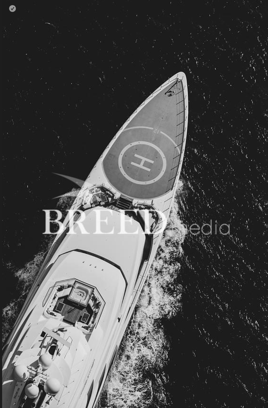 Aerial view of a superyacht cruising at sea with a large helipad on the bow, creating a white wake against dark blue water.