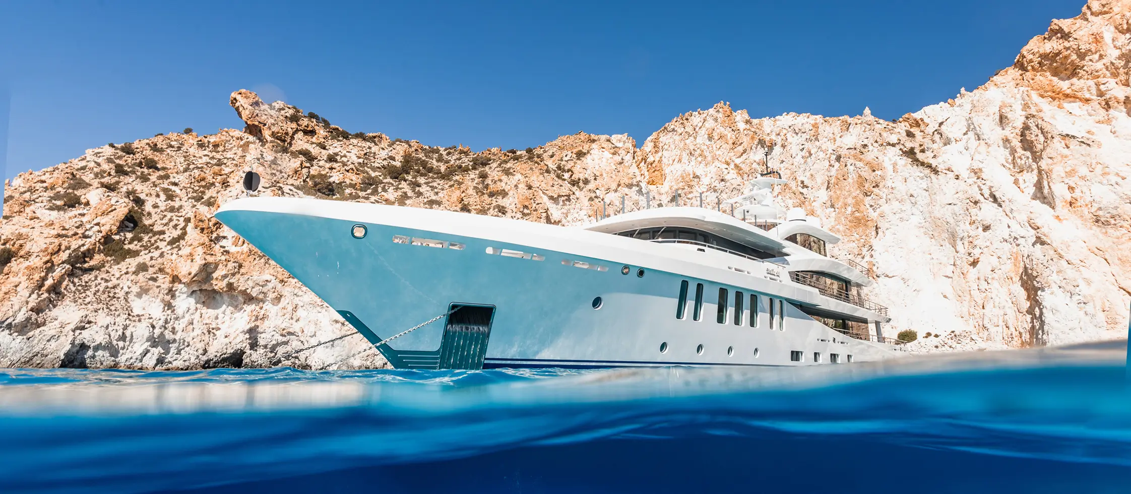 About us 1 Luxury superyacht anchored in clear blue water beside dramatic coastal cliffs, highlighting the vessel’s bow design and premium superyacht craftsmanship.