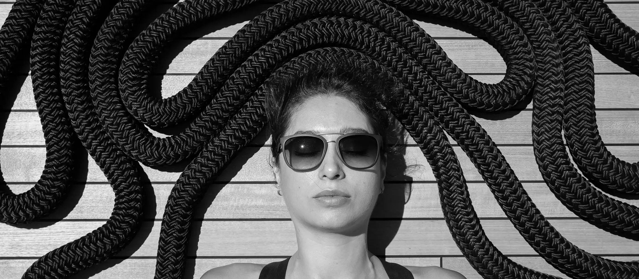 Woman relaxing on a yacht deck with large braided mooring ropes arranged around her head, wearing sunglasses and facing the sun.