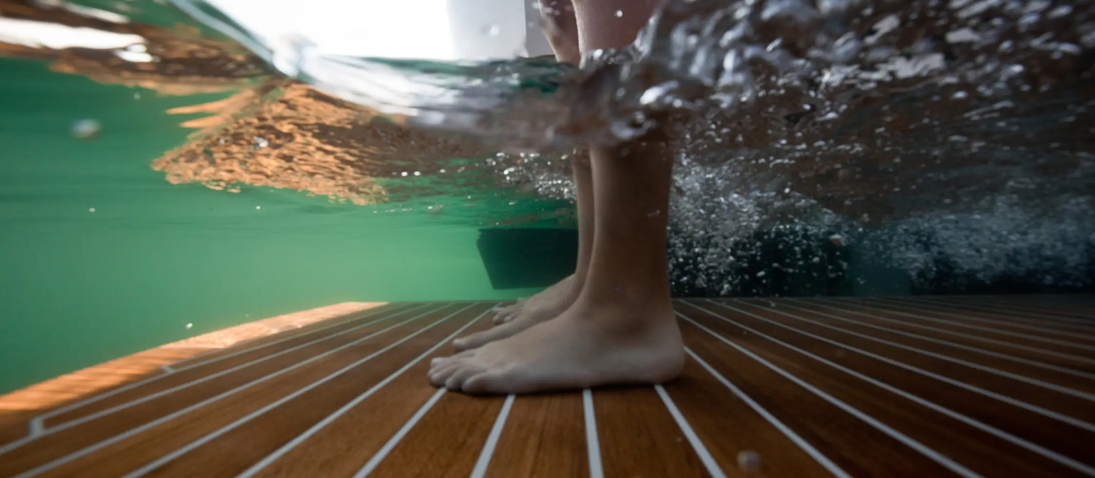 Underwater view of feet standing on a yacht swim platform with water splashing and sunlight filtering through