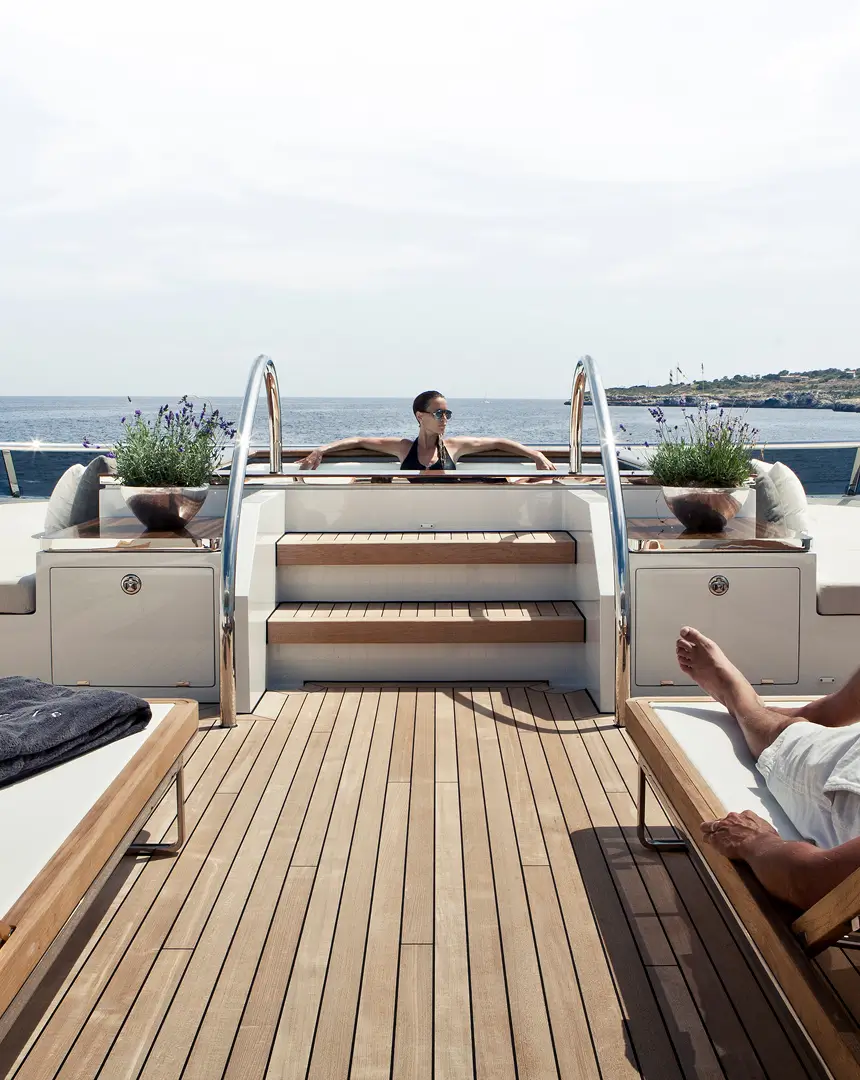Relaxed outdoor scene on a luxury yacht deck, with a person enjoying the sun near a raised seating area and others resting on loungers overlooking calm open water.
