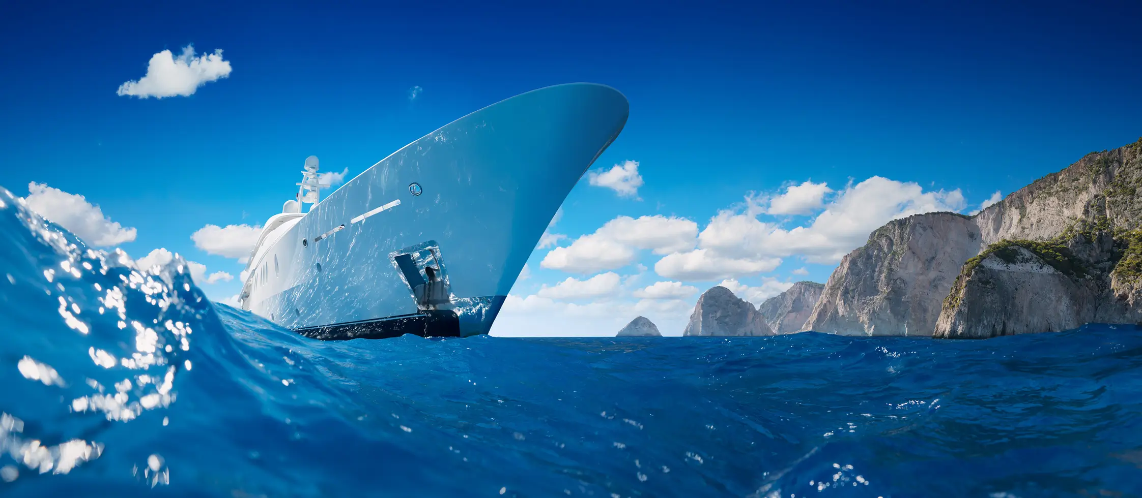 Yacht Refit and Conversion Projects 1 Superyacht cruising on open blue water near rocky coastline, photographed from sea level, illustrating luxury yacht ownership and the type of vessels involved in yacht purchase agreements and superyacht legal services.