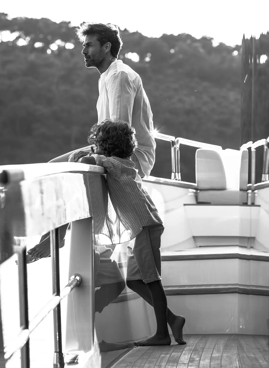 About us 5 Father and child standing on the deck of a luxury superyacht, leaning on the rail and looking out toward the sea during a calm, sunlit cruise.