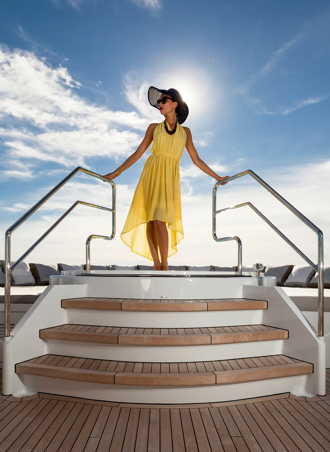 A woman in a flowing yellow dress standing on teak steps of a luxury superyacht, holding the railings under a bright sky, showcasing elegant yacht lifestyle and deck design.