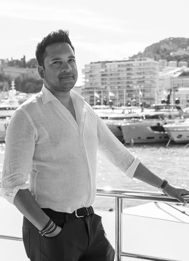 About us 6 Professional portrait of Alex Teji standing on a yacht deck overlooking a marina with boats and waterfront buildings in the background
