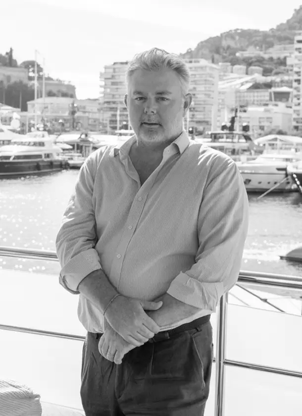 About us 7 Professional portrait of Antony Cowie, Co-Founder of Teji | Cowie and a maritime lawyer, standing on a yacht deck with a marina and waterfront buildings in the background.